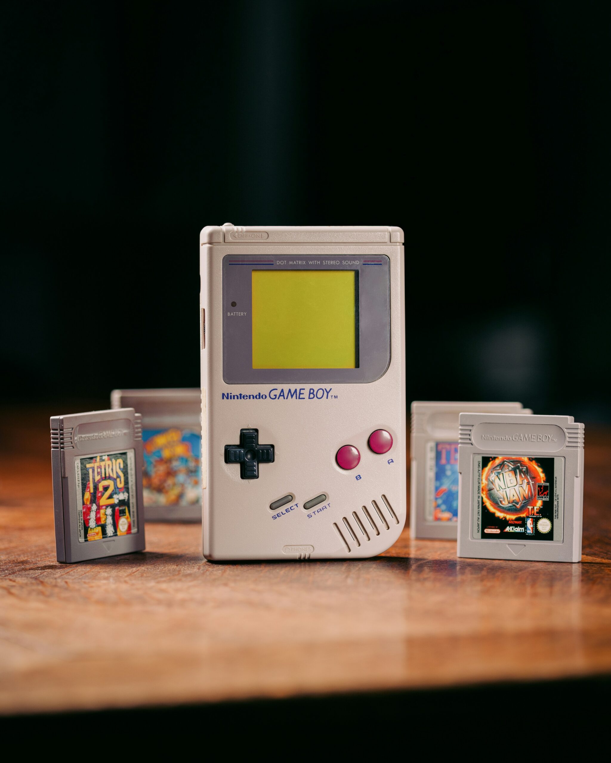 A Nintendo Game Boy standing upright on a wooden table, surrounded by several classic game cartridges including Tetris 2 and NBA Jam.