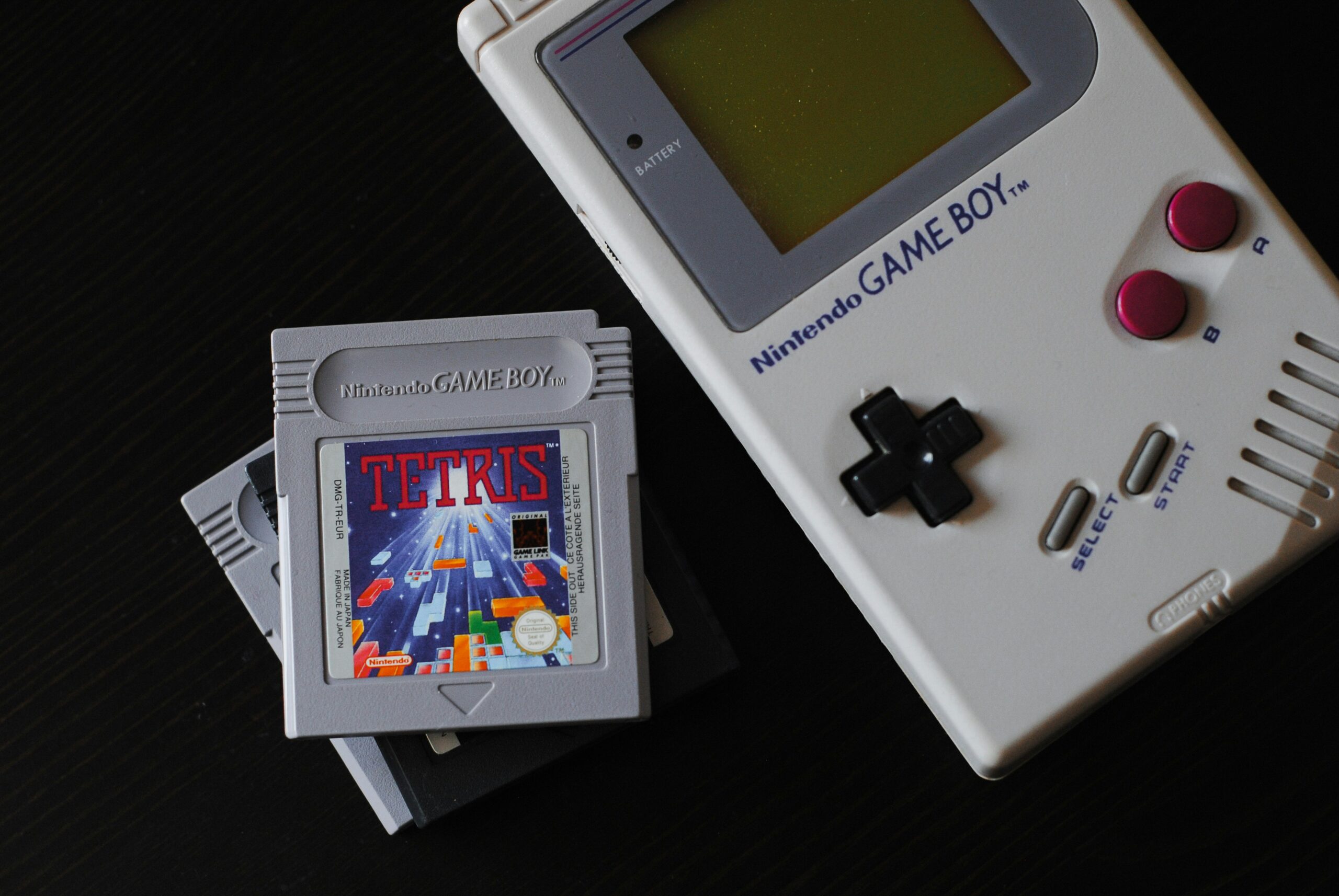 A close-up view of a classic Nintendo Game Boy next to a stack of cartridges, with a Tetris game cart displayed on top.